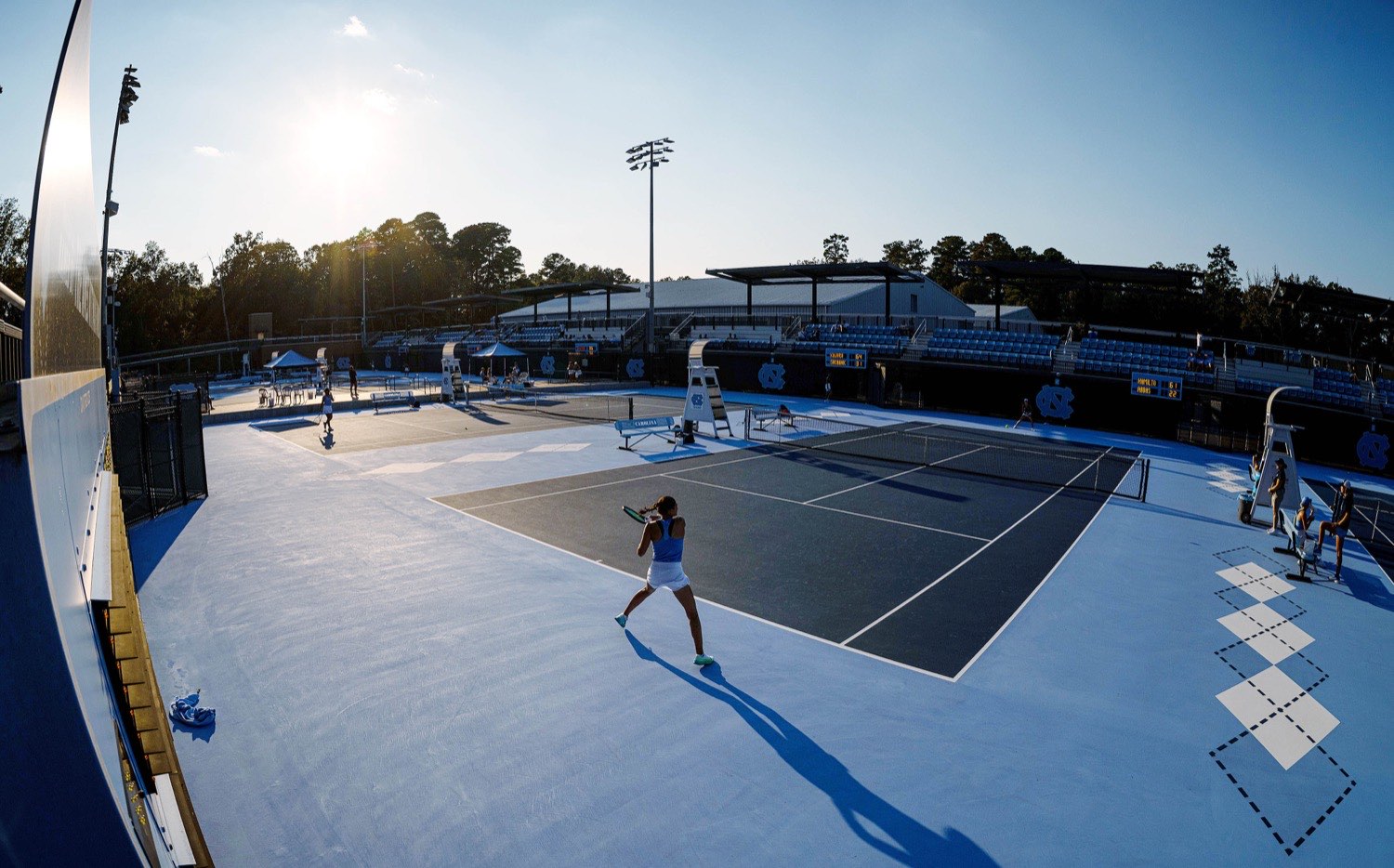A Carolina player striking a forehand during a match at the Chewning Tennis Center, with play under way on the courts behind her and live scoreboards on the back wall.